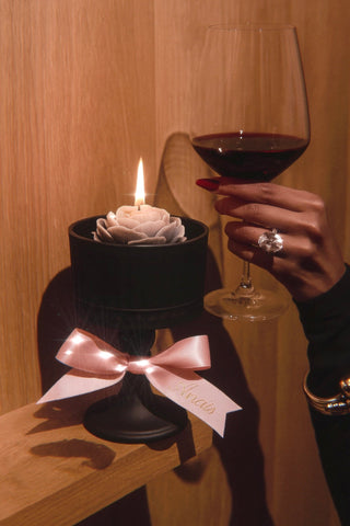 Candle in a wine glass holder with a pink bow, held by a hand with a ring, against a wooden background.