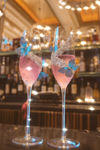 Two pink cocktails with butterfly decorations in decorative glasses on a bar counter.
