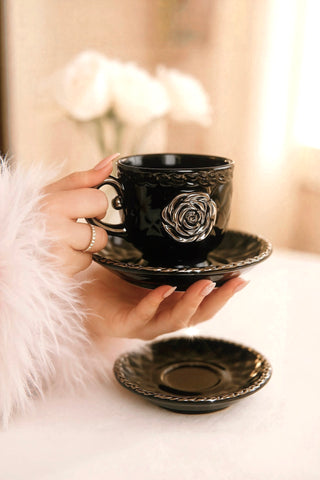 Black ornate teacup and saucer held by a hand with a blurred floral background..