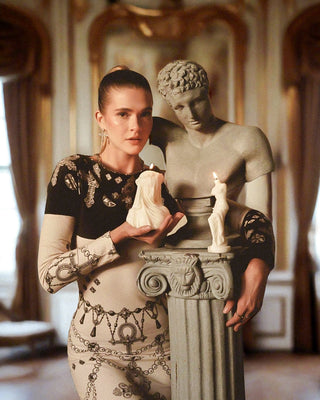 A woman holding an 'Amour, Anais' candle next to a Roman and Greek statue.