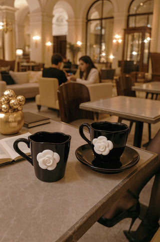 Two black mugs with white floral designs on a table in a cozy café setting.