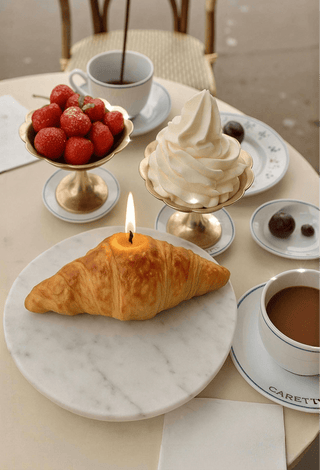 A lit French Croissant Candle next to pastries and coffee on a Paris sidewalk during brunch.
