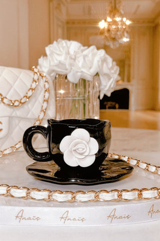 Black teacup with white floral design on a saucer, placed on a decorative tray with 'Anais' branding.
