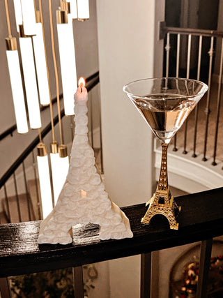 Rosy Eiffel Tower Candle in White, atop a banister, next to an Eiffel Tower glass cup.