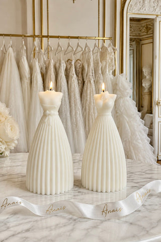 Two white pleated pillar candles shaped like wedding dresses sit on a marble table in front of a rack of white bridal dresses, with a white ribbon printed with the brand name Anais.