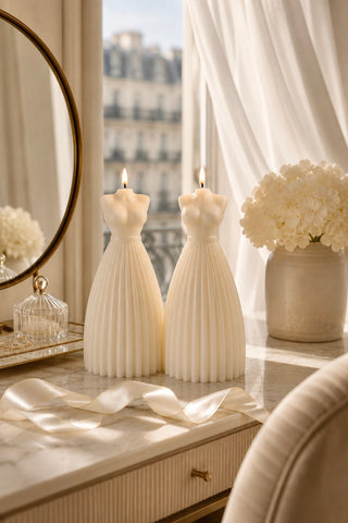 Two cream pleated dress-shaped candles on a marble vanity table, styled with a mirror, ribbon, and a vase of white flowers in a softly lit room.