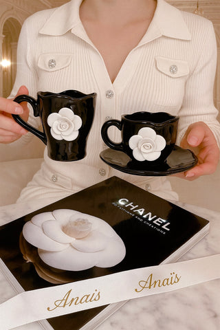 Person holding two black mugs with white flower designs and a Chanel box on a marble surface.
