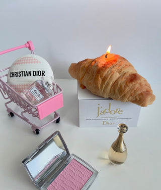 A lit French Croissant Candle next to luxurious makeup items.