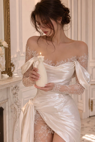 A woman in a white satin off-the-shoulder lingerie dress with lace sleeves holds a lit white candle in an ornate white room.
