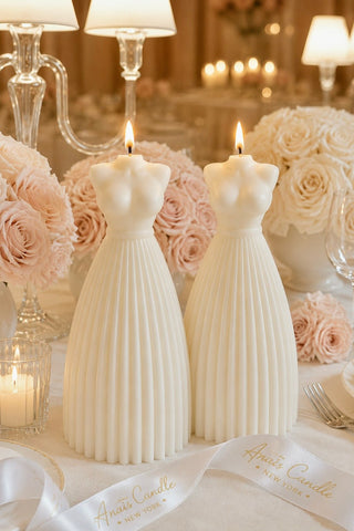 Two white dress-shaped taper candles with lit flames, styled on a table with blush pink roses, glass candleholders. 