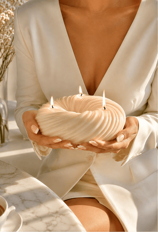 A person in a cream satin blazer holds a sculptural cream-colored candle with multiple wicks in their hands, set in a warm, sunlit interior next to a marble table and a cup of coffee.