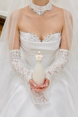 A bride in a white lace wedding dress and veil holds a white ribbed candle shaped like a small mannequin torso. The dress has a lace choker and long lace sleeves, photographed against a softly blurred indoor background.