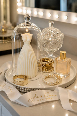 A styled vanity scene with a white dress-shaped candle under a clear glass cloche, a pearl strand, gold rings, a perfume bottle, and a decorative tray with a branded ribbon reading Anais Carolina New York.